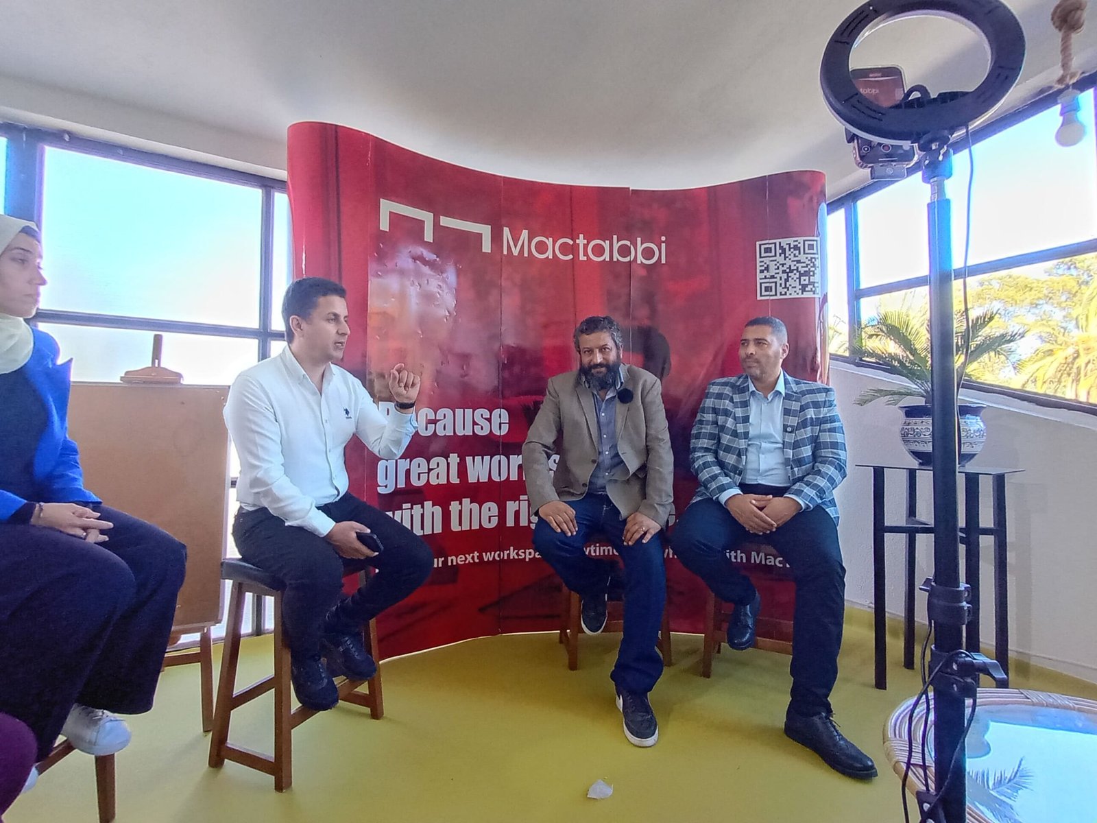 Trois hommes discutant devant un fond rouge avec le logo de Mactabbi lors d'un événement.